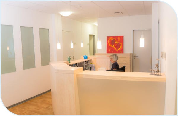 Ein Empfangsbereich in einer Arztpraxis mit einem hellen Holzschalter und einer sitzenden Person im Hintergrund.