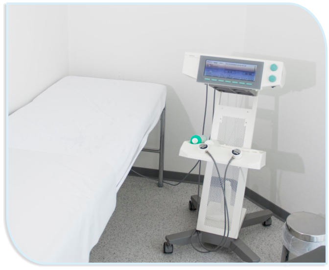 Ein leerer, sauberer Behandlungsraum mit einer Untersuchungsliege auf der linken Seite und einem modernen Ultraschalltherapiegerät auf einem Rollständer auf der rechten Seite.