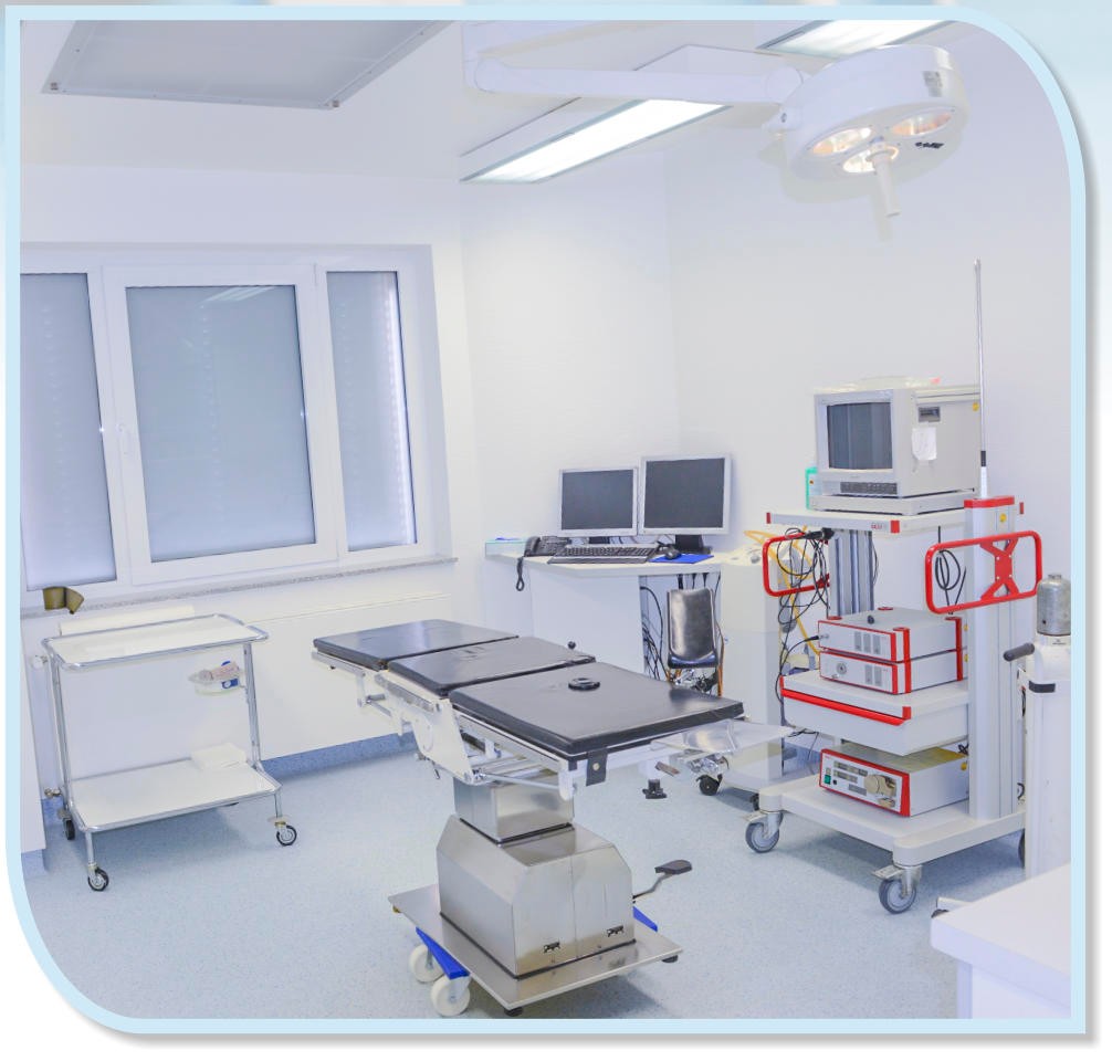 Heller und moderner Operationssaal mit einem Operationstisch in der Mitte, einer Operationsleuchte an der Decke und verschiedenen medizinischen Geräten auf Rollwagen.